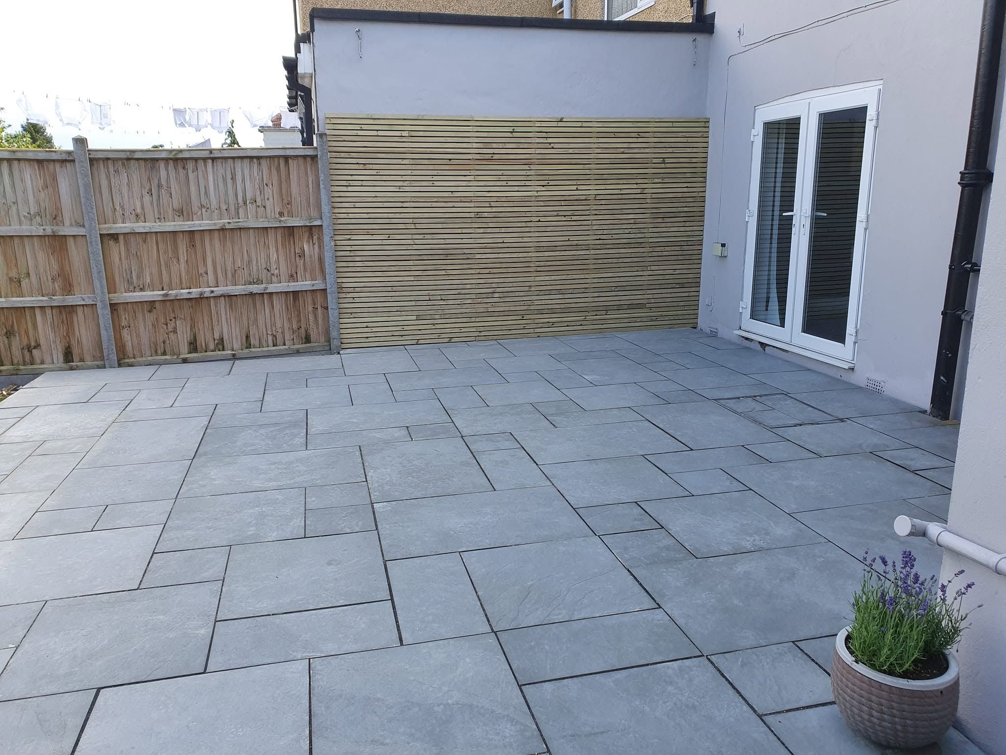 New grey stone patio laid in mixed-size slabs beside French doors, with a slatted timber screen and potted lavender