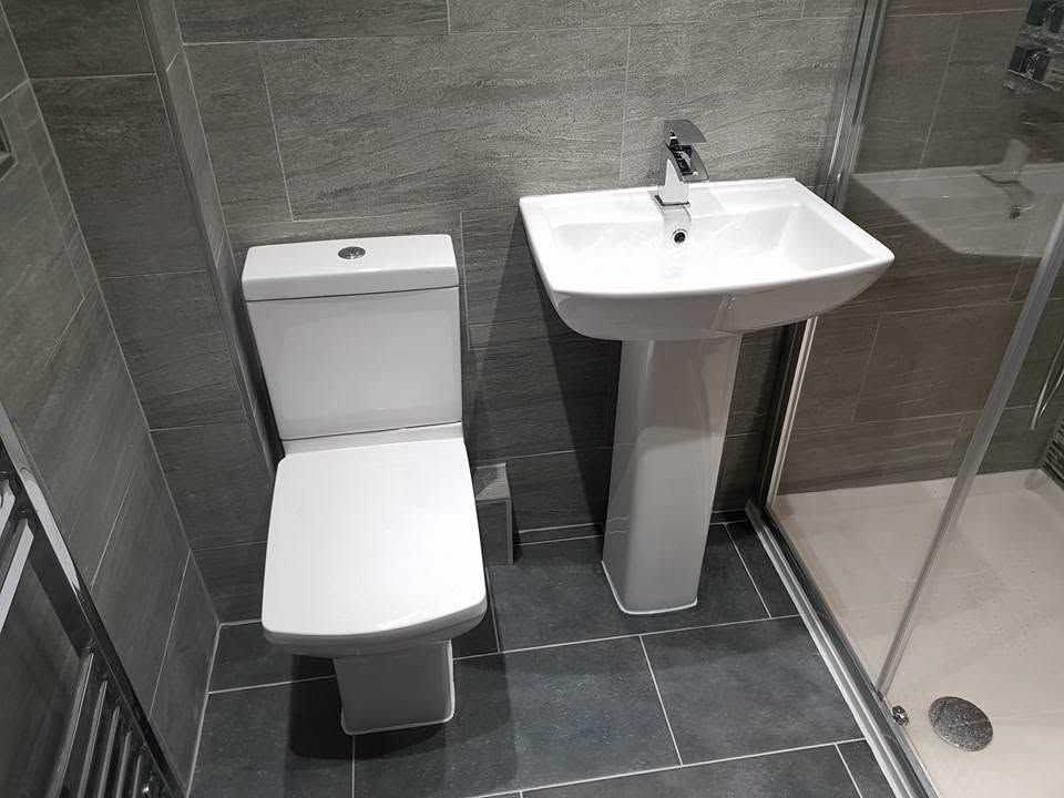 Square white toilet and pedestal basin against grey wood-effect wall tiles with dark floor tiles and a shower tray