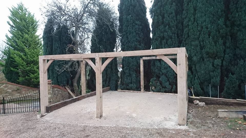 Green oak frame with curved braces standing on a prepared gravel base in front of tall conifers