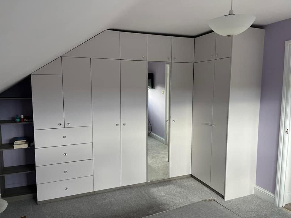 Light grey fitted wardrobes shaped to a sloping ceiling with drawers, mirrored door and open side shelving