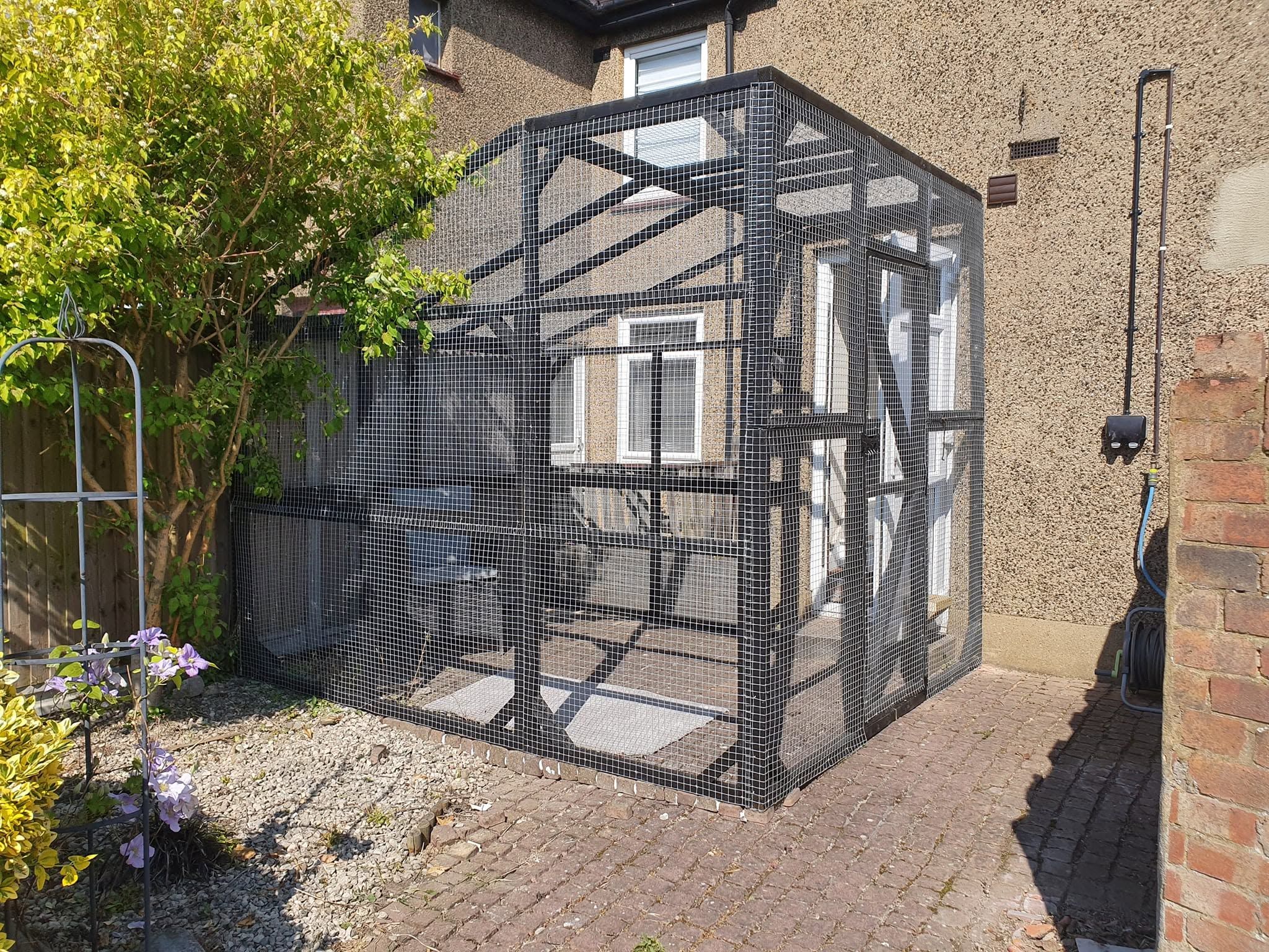 Black-painted timber-framed mesh enclosure with braced frame and door, built against the side of a house